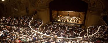 The interior of merrill auditorium from just above the back of the balcony with a full crowd, and a full symphony on stage.