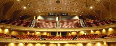A photo of the inside of Merrill Auditorium as viewed from the stage. Three levels of seating with maroon chairs and house lights.