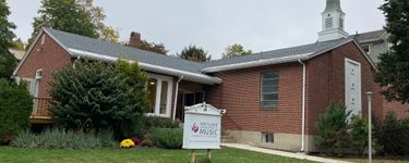 The exterior of the portland conservatory of music. It is a small converted brick church with a small steeple to the right on the front part of the building. A sign on the lawn has the portland conservatory of music logo on it. a large green bush covers the left side of the building.
