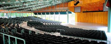 The Old orchard beach seaside pavilion from the back of the seating on the far right of the amphitheater. The seating is black with 1970s green railings and metal structures above the seating. The stage itself is white, with a bright finished wood shell behind and above the stage.